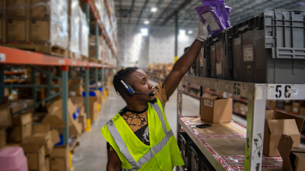 A warehouse worker wears a headset and picks an item from an inventory shelf.